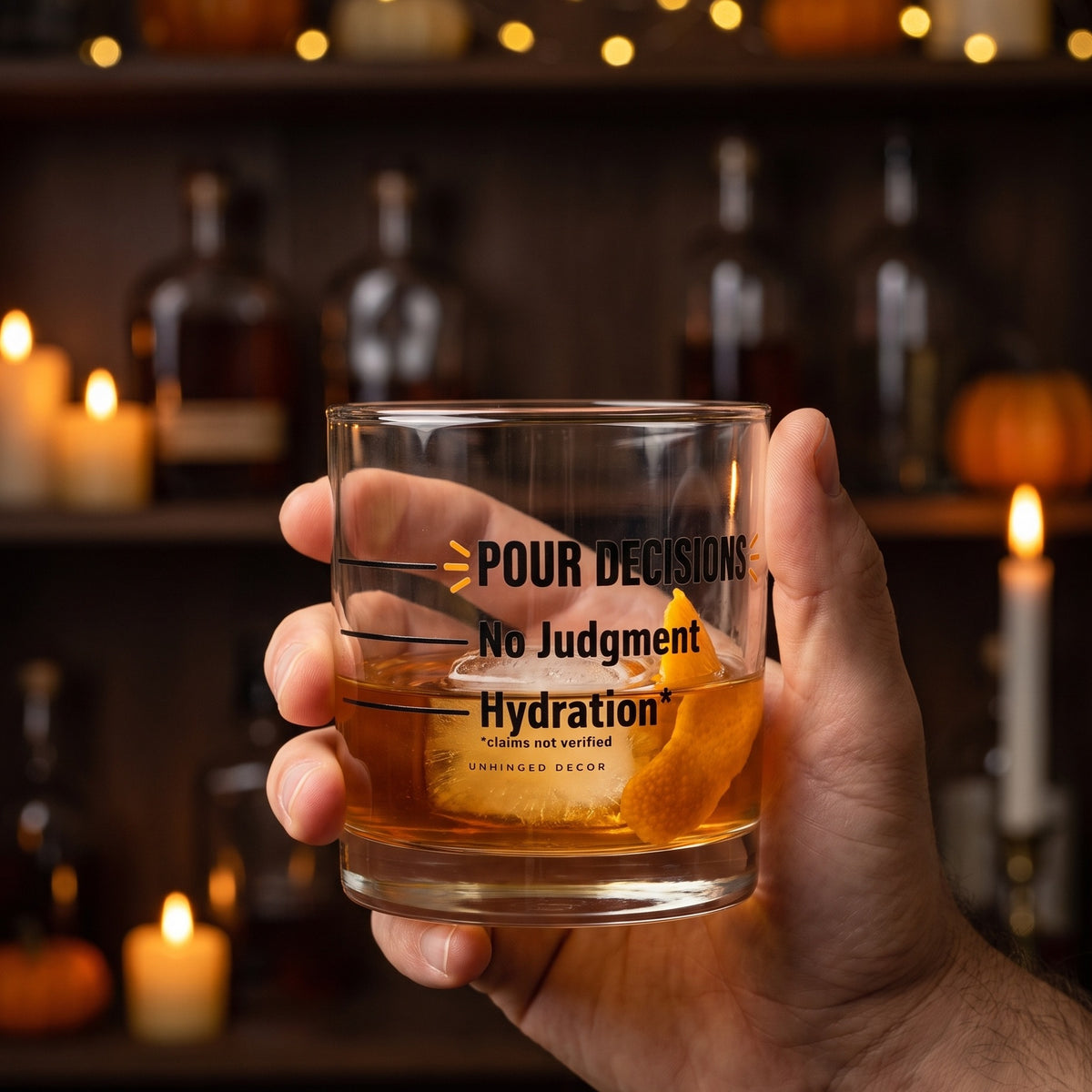 Hand holding the Pour Decision rocks glass 'Hydration* / No Judgment' with whiskey, ice and orange peel in a candlelit fall home bar, Unhinged Decor