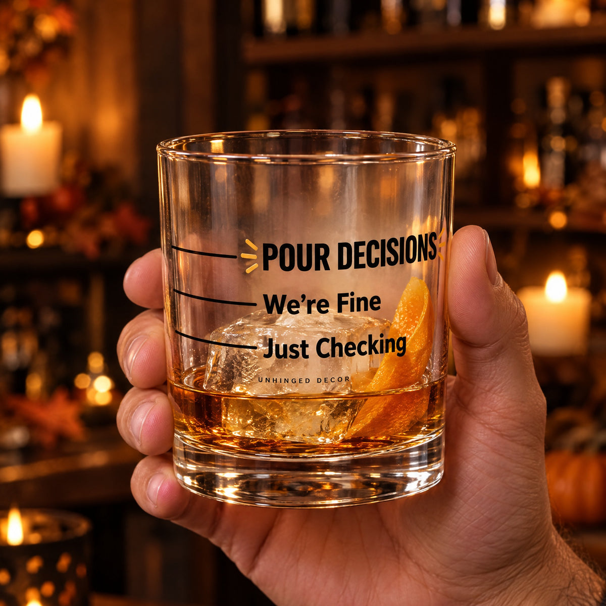 Hand holding the Pour Decision rocks glass 'Just Checking / We’re Fine' with whiskey, ice and orange peel in a candlelit fall home bar, Unhinged Decor